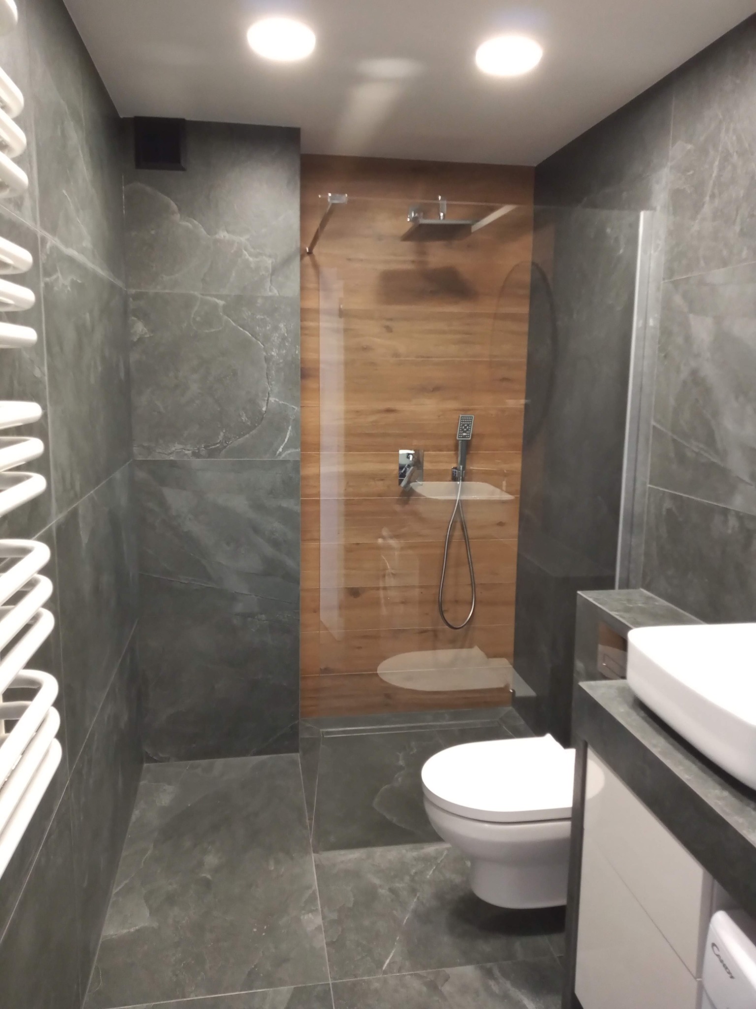 Nowoczesna łazienka z kabiną prysznicową typu walk-in z drewnopodobną ścianą, szare płytki imitujące kamień na ścianach i podłodze, biała toaleta wisząca i umywalka nablatowa.