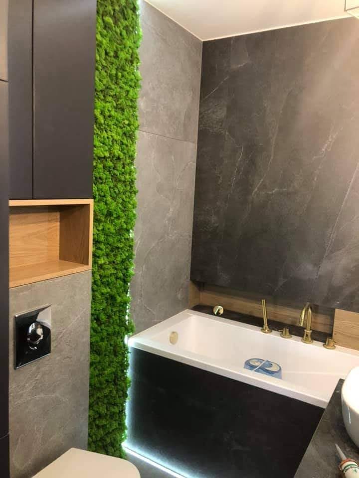 Nowoczesna łazienka z pionową ścianą z mchu, szare płytki imitujące marmur, biała wanna z czarną obudową i złotą armaturą, oraz podświetleniem LED u dołu, obok toaleta.