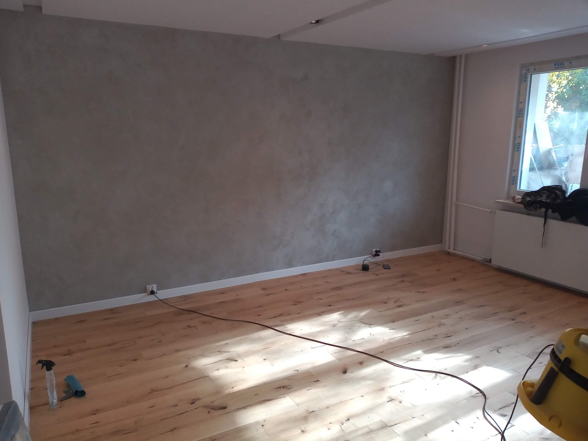 Świeżo wykończony pokój z panelami podłogowymi i dekoracyjną szarą ścianą, widoczne gniazdka elektryczne przy listwach przypodłogowych, narzędzia i odkurzacz w tle.