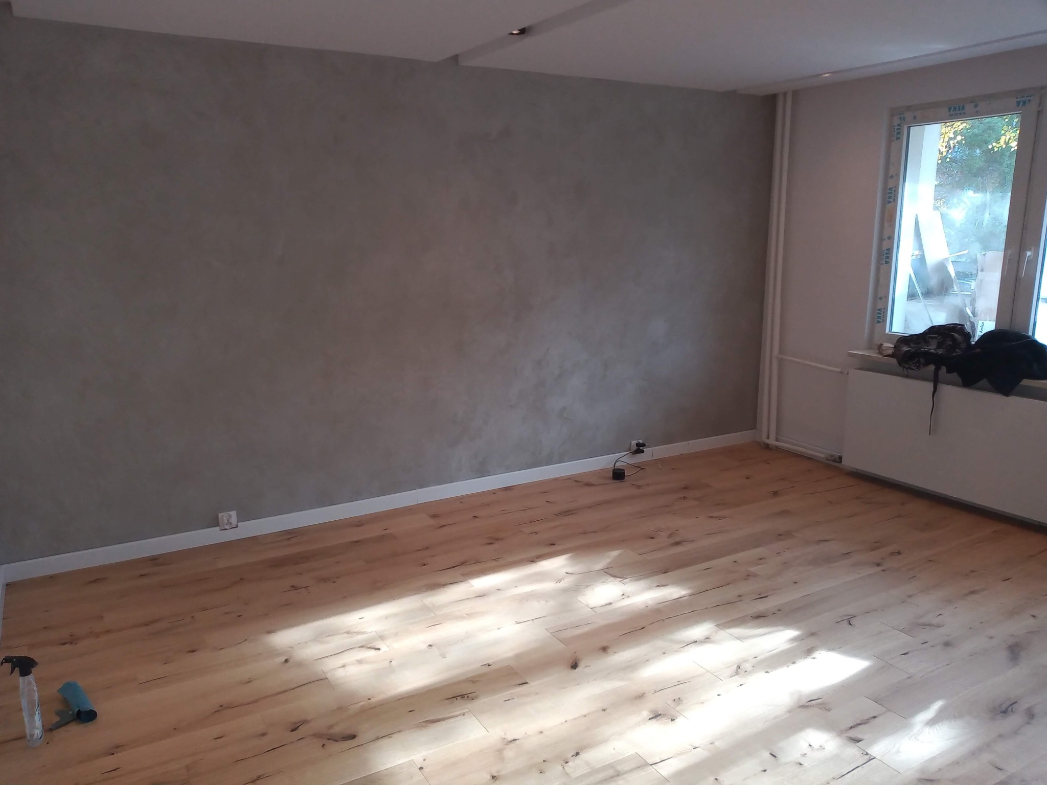 Wykończone wnętrze pokoju z parkietem o jasnym odcieniu drewna, ściana w odcieniach szarości z widoczną fakturą, biała listwa przypodłogowa, okno z widokiem na zewnątrz, grzejnik pod oknem,...