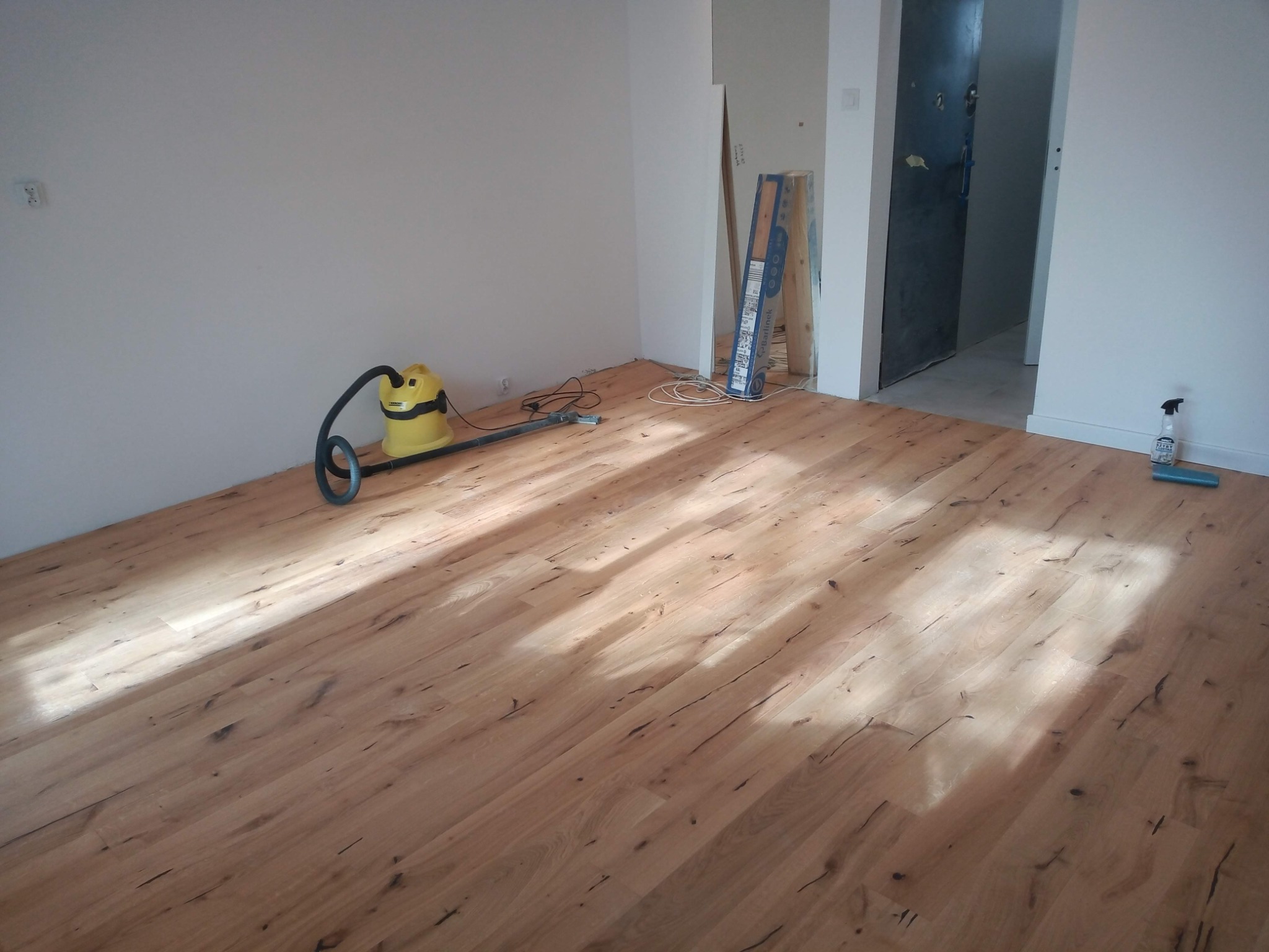 Nowo ułożona podłoga z paneli w jasnym odcieniu drewna, z widocznym odkurzaczem przemysłowym Karcher, opakowaniem paneli Bartlinek i butelką środka czyszczącego, sugerująca etap sprzątania po montażu.