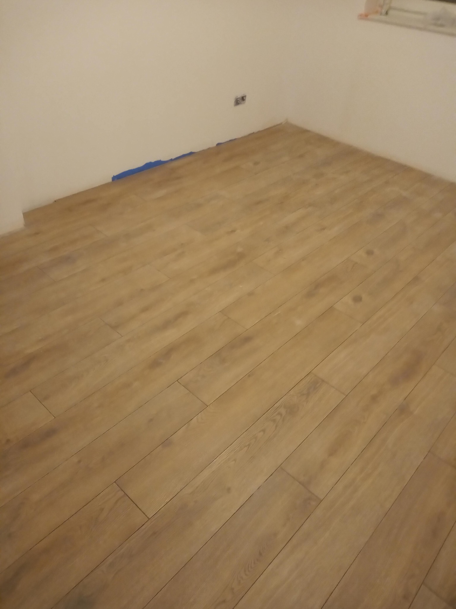 Ułożone panele podłogowe w rogu pokoju, zabezpieczone niebieską taśmą malarską przy ścianie, widoczne gniazdko elektryczne.