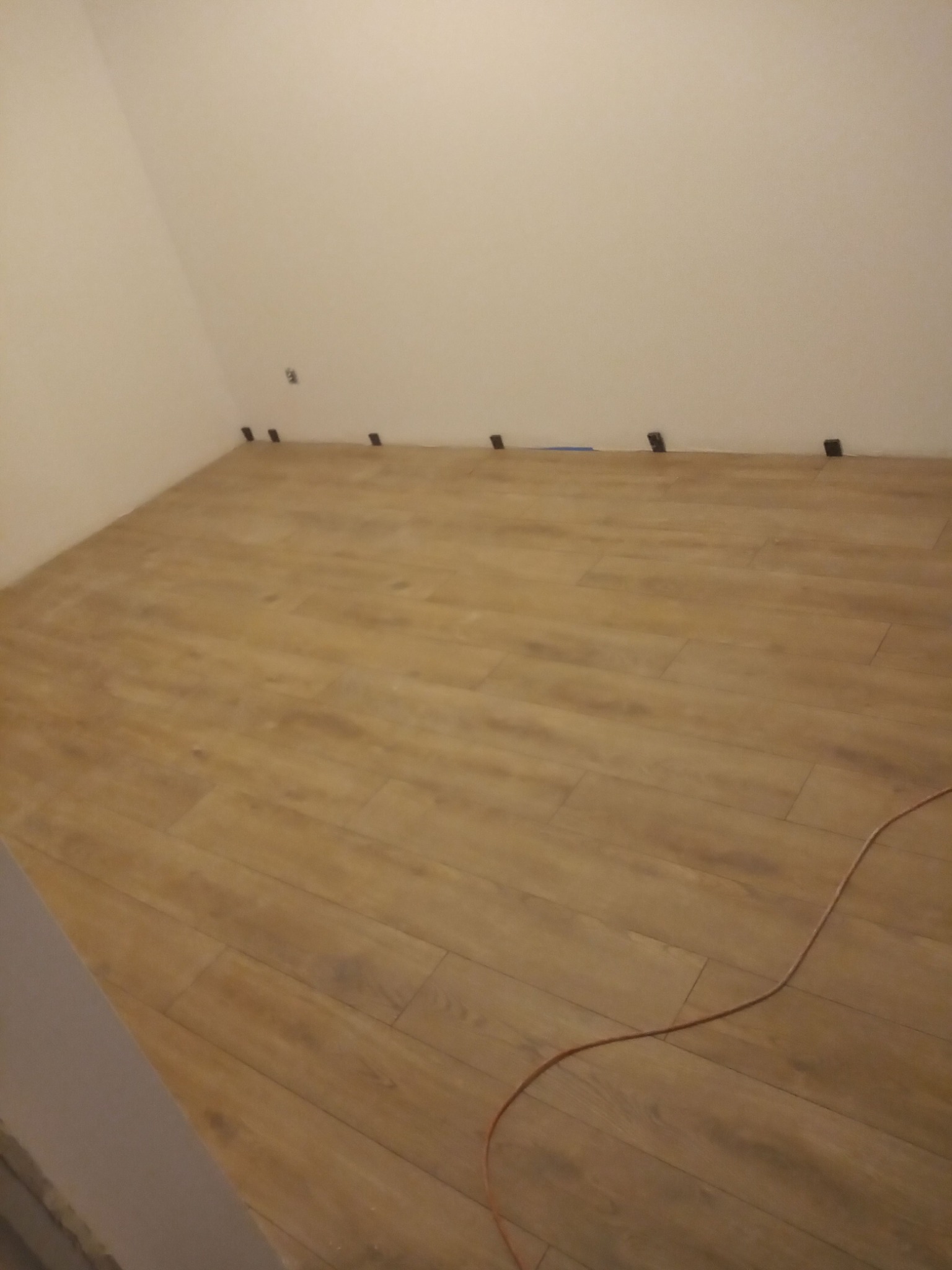 Fragment pokoju z ułożonymi panelami podłogowymi w odcieniach brązu, widoczne czarne kliny dystansowe przy ścianie i pomarańczowy kabel na podłodze.