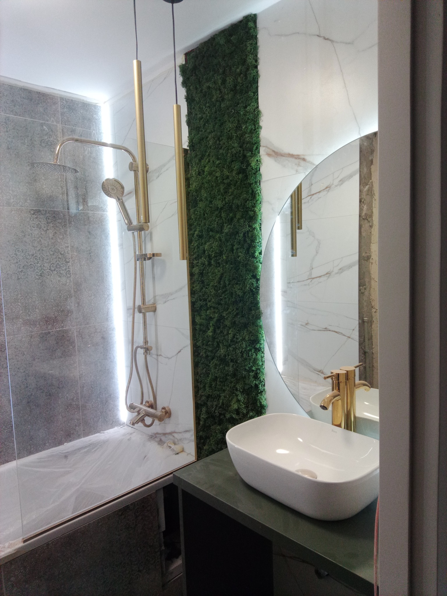 Nowoczesna łazienka z zielonym panelem z mchu, złotą armaturą i owalną umywalką nablatową, widoczny prysznic za szklaną ścianką.
