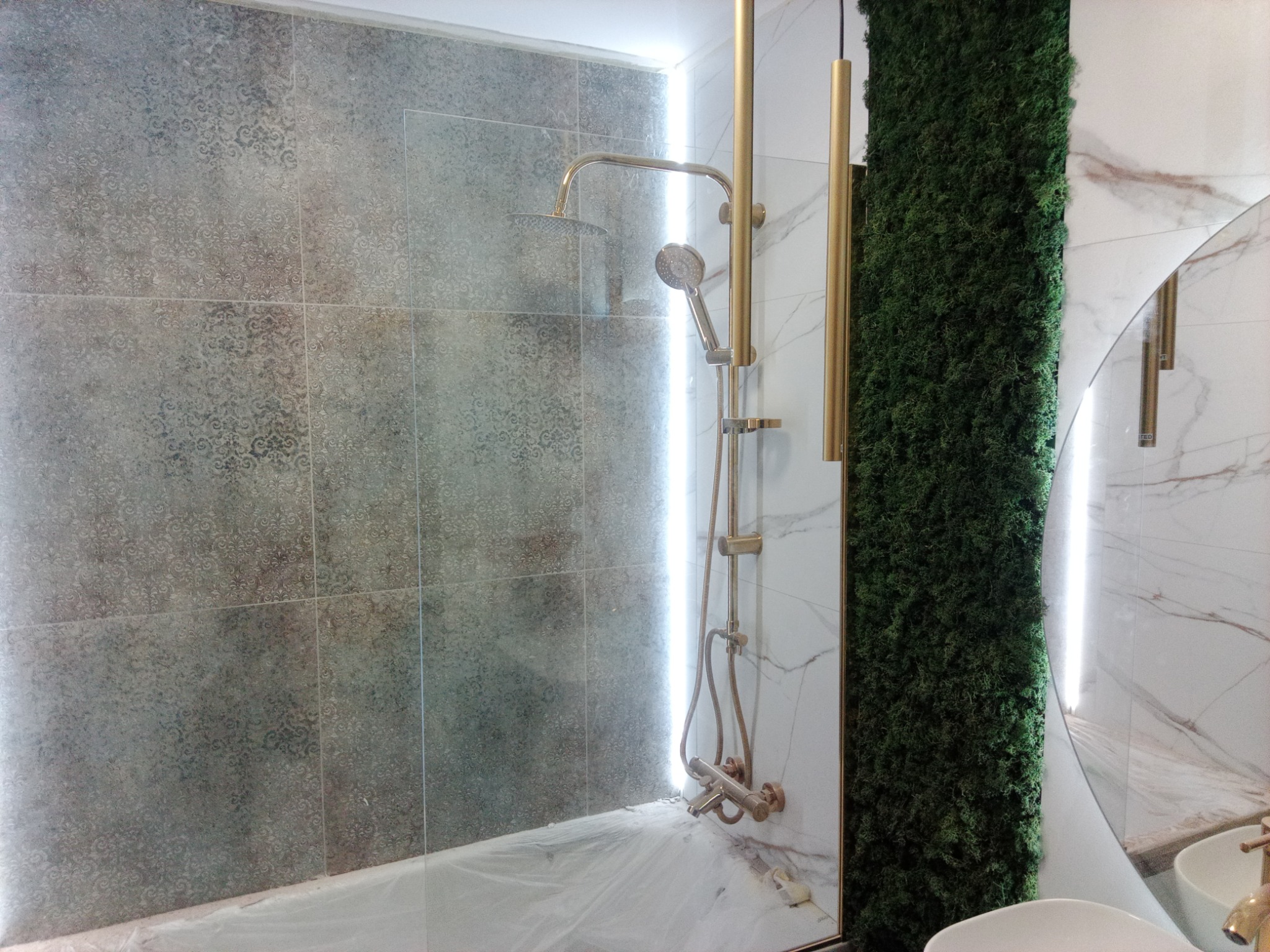 Nowoczesna łazienka z kabiną prysznicową typu walk-in, wykończona płytkami w odcieniach szarości z delikatnym wzorem, złotą armaturą i panelem z mchu dekoracyjnego.