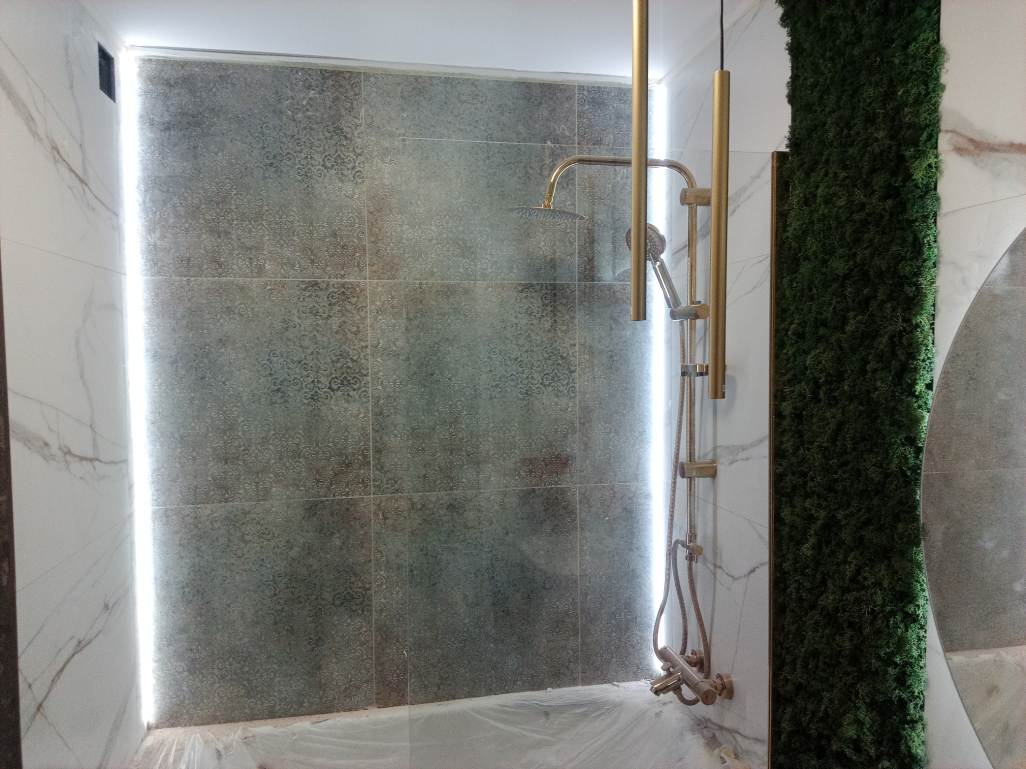 Nowoczesna kabina prysznicowa z szaro-wzorzystymi płytkami, złotą armaturą i pionową ścianą z mchu dekoracyjnego, zabezpieczona folią ochronną na podłodze.