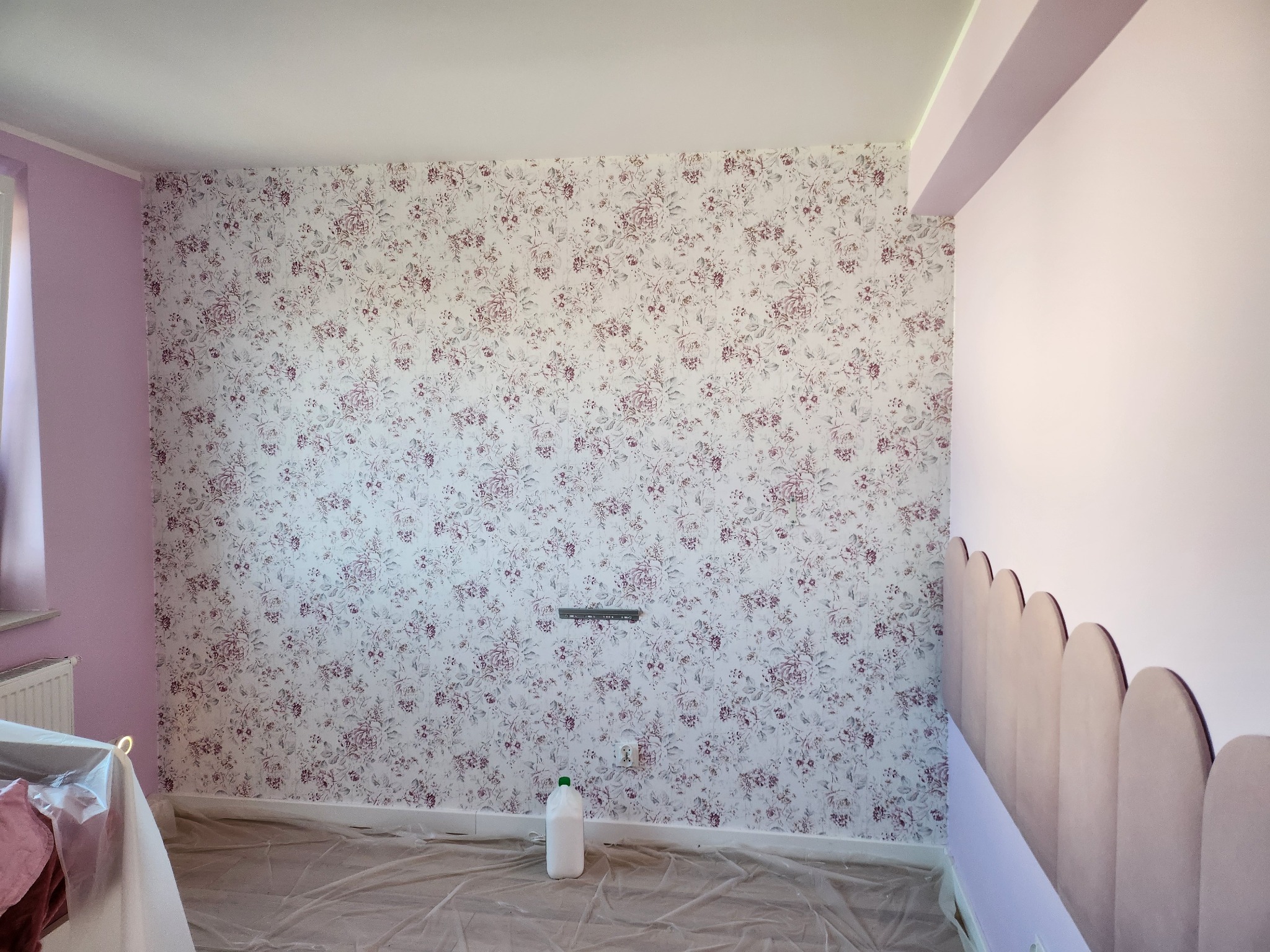 Pokój z jedną ścianą w kwiatowy wzór i drugą pomalowaną na liliowo, zabezpieczony folią malarską, widoczna butelka z płynem i poziomica na ścianie.