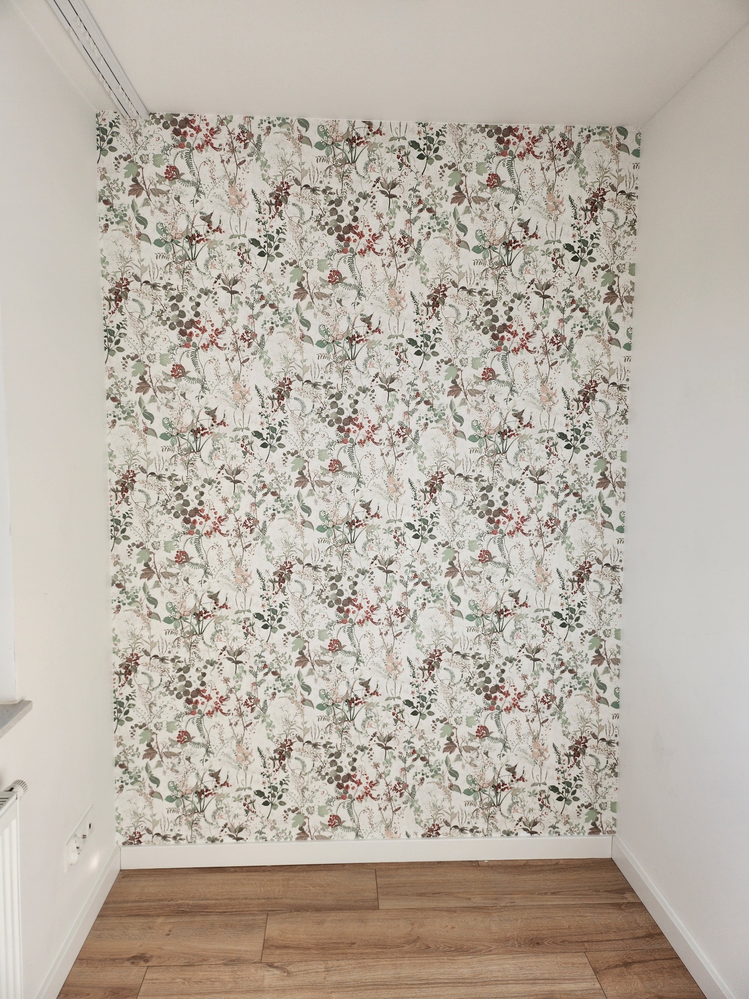 Ściana pokoju wykończona tapetą w kwiatowe wzory, z widocznymi panelami podłogowymi i białymi ścianami bocznymi.
