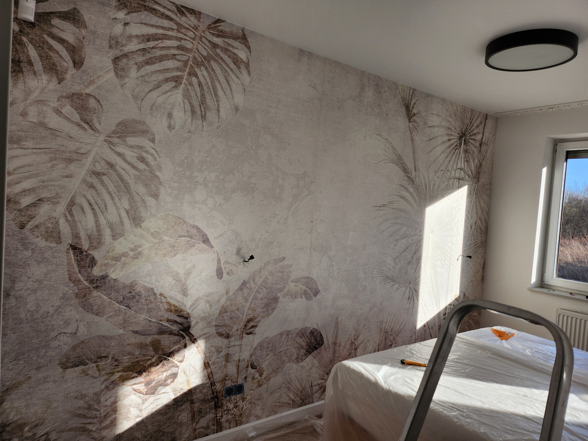 Ściana po malowaniu z motywem roślin tropikalnych, zabezpieczona folią malarską, widoczna drabina malarska, oświetlona silnym światłem słonecznym padającym z okna.