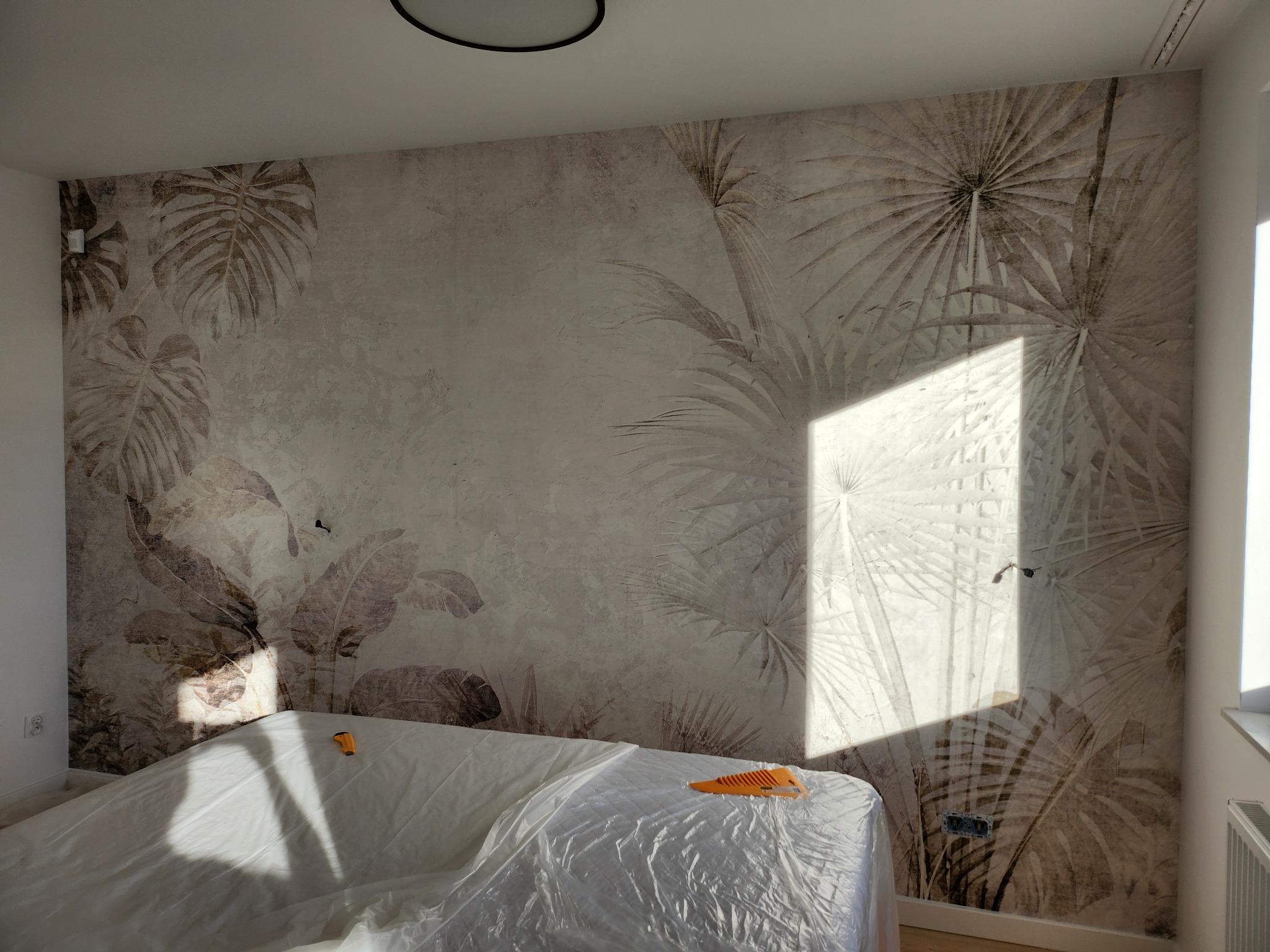 Ściana z fototapetą w motywy roślinne, zabezpieczone meble folią malarską, widoczny fragment narzędzia do tapetowania.