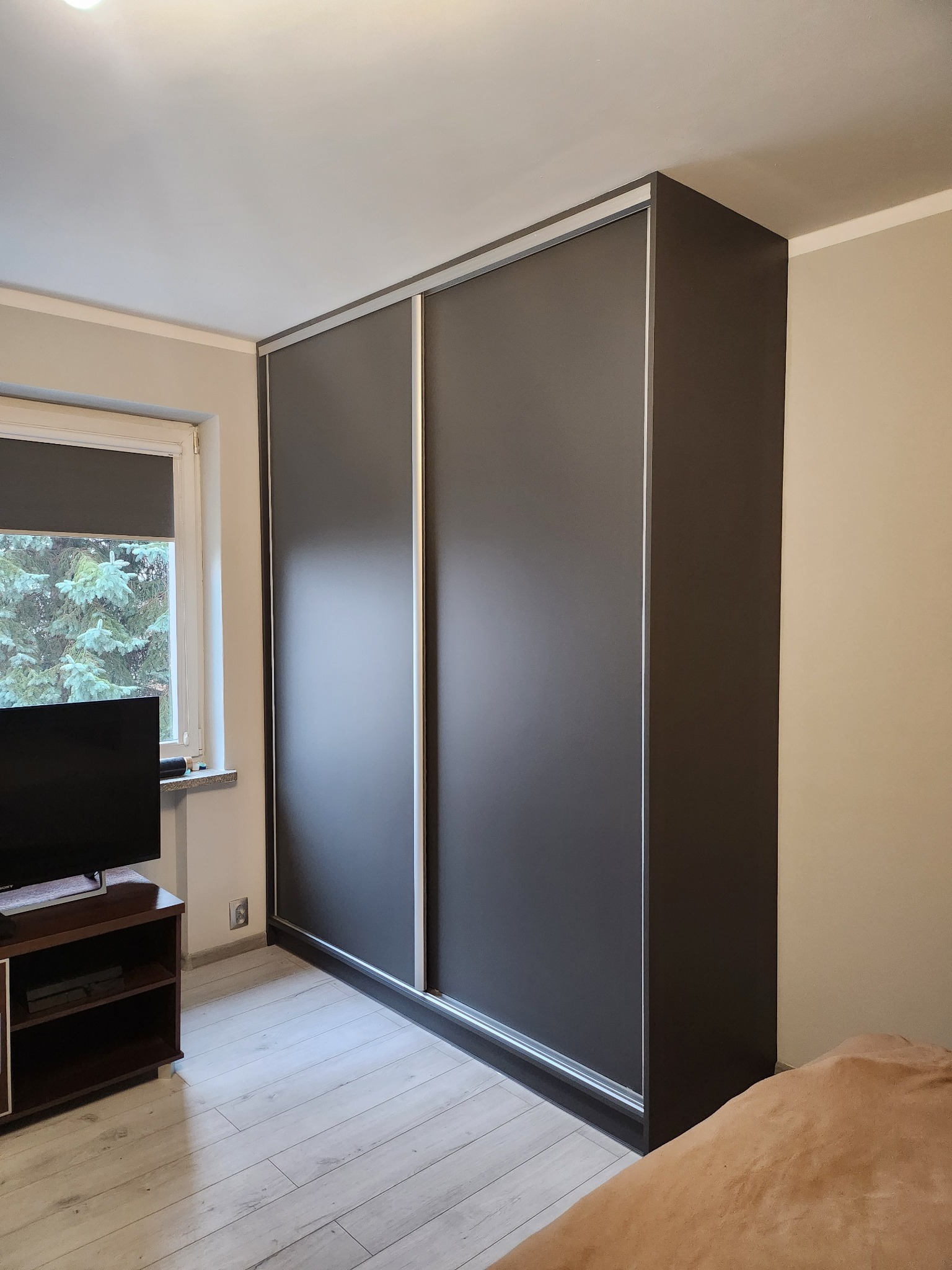 Ciemnoszara szafa przesuwna z srebrnymi obramowaniami, widoczna w rogu pokoju z jasną podłogą z paneli i telewizorem na szafce RTV obok okna.