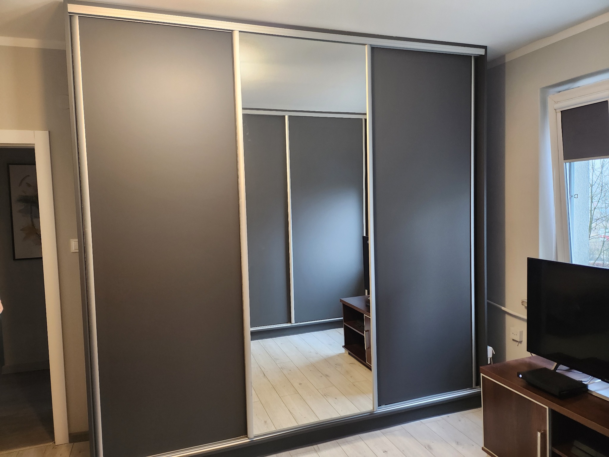 Szara szafa przesuwna z lustrzanymi drzwiami, widoczne wnętrze szafy i odbicie podłogi, obok telewizor na szafce.