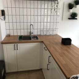 “Ręce Mistrza” usługi remontowe Anton Velychko - Białe szafki kuchenne z drewnianym blatem, chromowany zlew i bateria, białe kafelki z czarną fugą na ścianie, zioła w doniczkach na półce i ścianie.