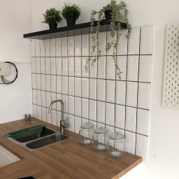 “Ręce Mistrza” usługi remontowe Anton Velychko - Nowoczesna kuchnia z drewnianym blatem, białymi płytkami typu metro z czarną fugą, metalowym zlewem i baterią, oraz dekoracyjnymi słoikami i roślinami doniczkowymi na półce i tablicy perforowanej.