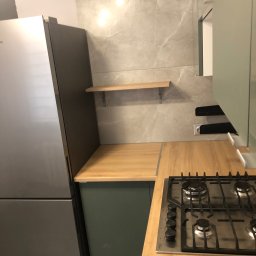 “Ręce Mistrza” usługi remontowe Anton Velychko - Fragment kuchni z szarą lodówką Samsung, blatem z płyty wiórowej, czteropalnikową płytą gazową i półką na ścianie wyłożonej płytkami imitującymi marmur.