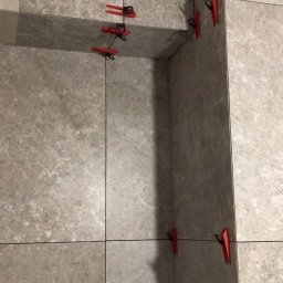 “Ręce Mistrza” usługi remontowe Anton Velychko - Szare płytki ścienne z systemem poziomowania płytek, czerwone klipsy i czarne kliny utrzymują równe odstępy.