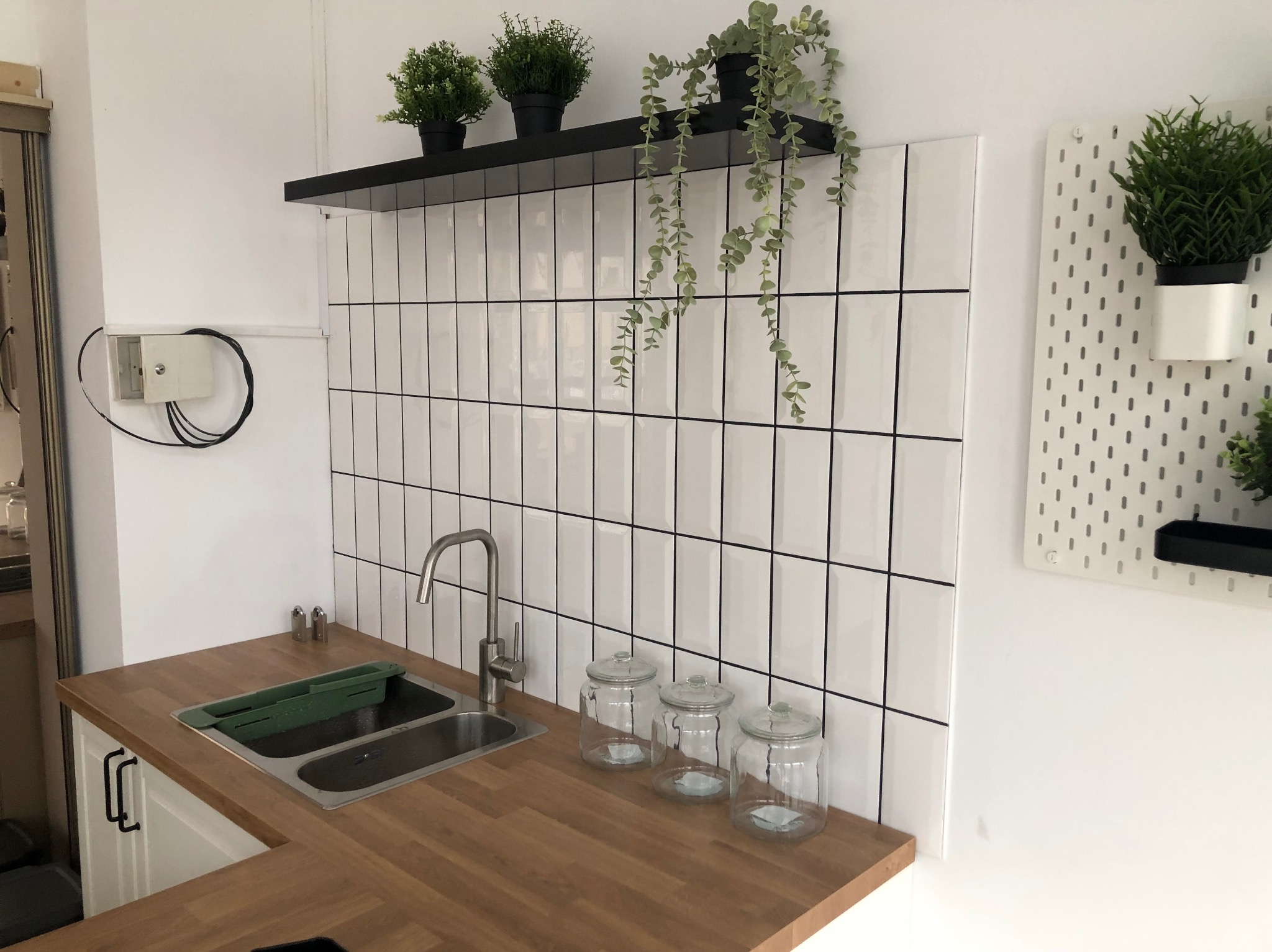 Nowoczesna kuchnia z drewnianym blatem, białymi płytkami typu metro z czarną fugą, metalowym zlewem i baterią, oraz dekoracyjnymi słoikami i roślinami doniczkowymi na półce i tablicy perforowanej.