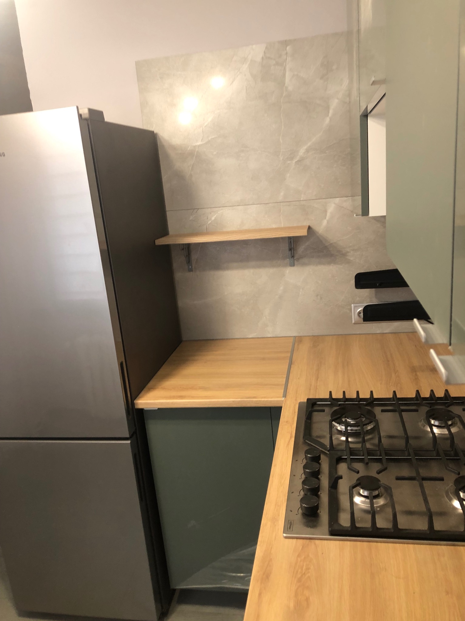 Fragment kuchni z szarą lodówką Samsung, blatem z płyty wiórowej, czteropalnikową płytą gazową i półką na ścianie wyłożonej płytkami imitującymi marmur.