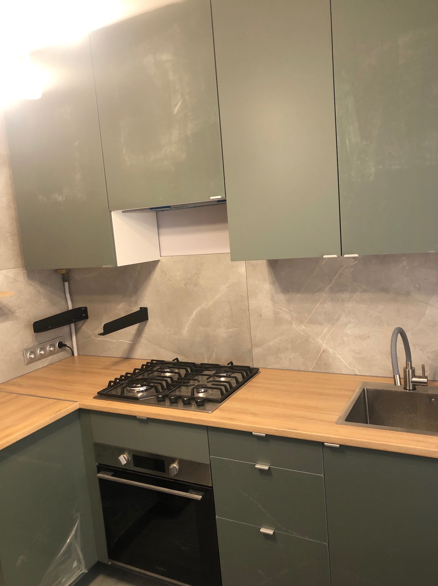 Nowoczesna kuchnia z frontami w odcieniach zieleni, drewniany blat, płyta gazowa i marmurowe płytki na ścianie.