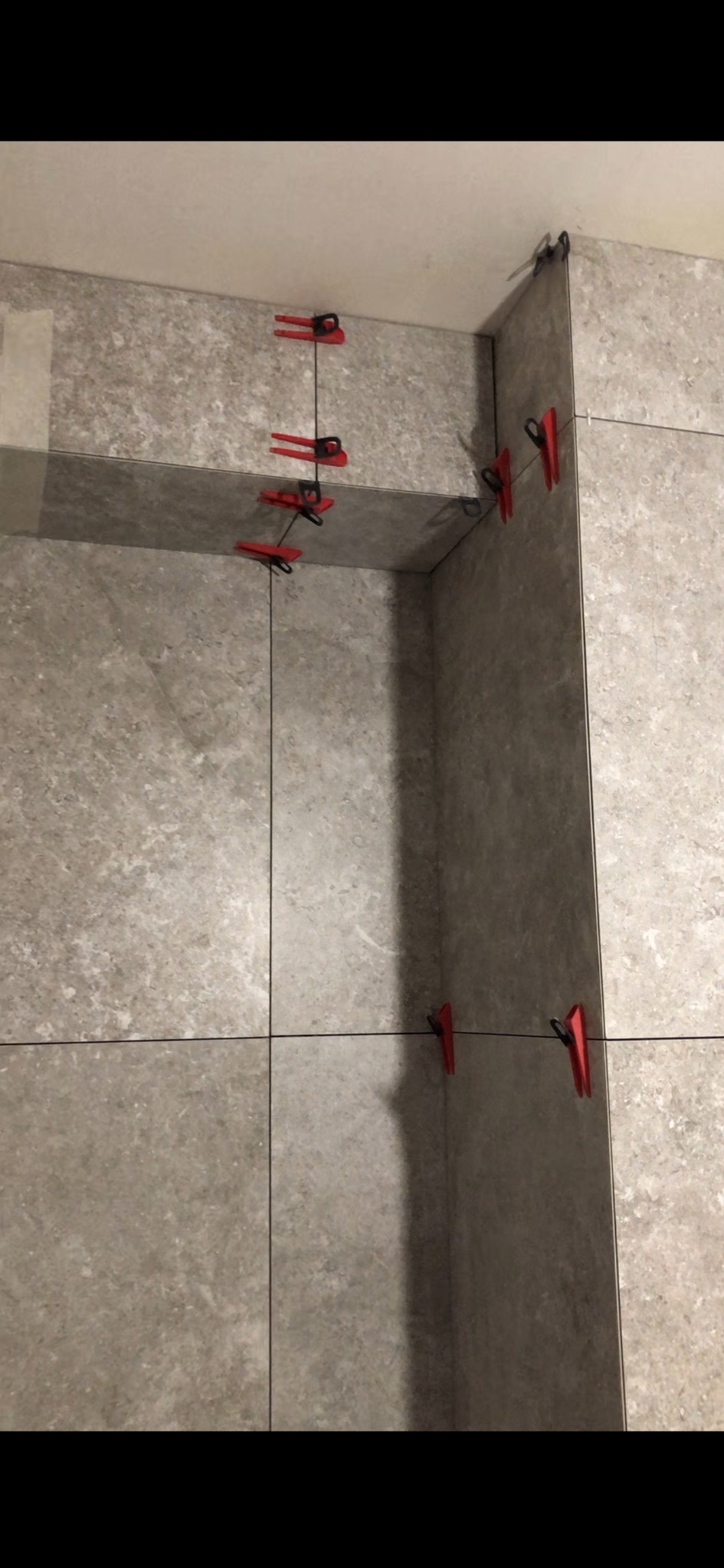 Szare płytki ścienne z systemem poziomowania płytek, czerwone klipsy i czarne kliny utrzymują równe odstępy.