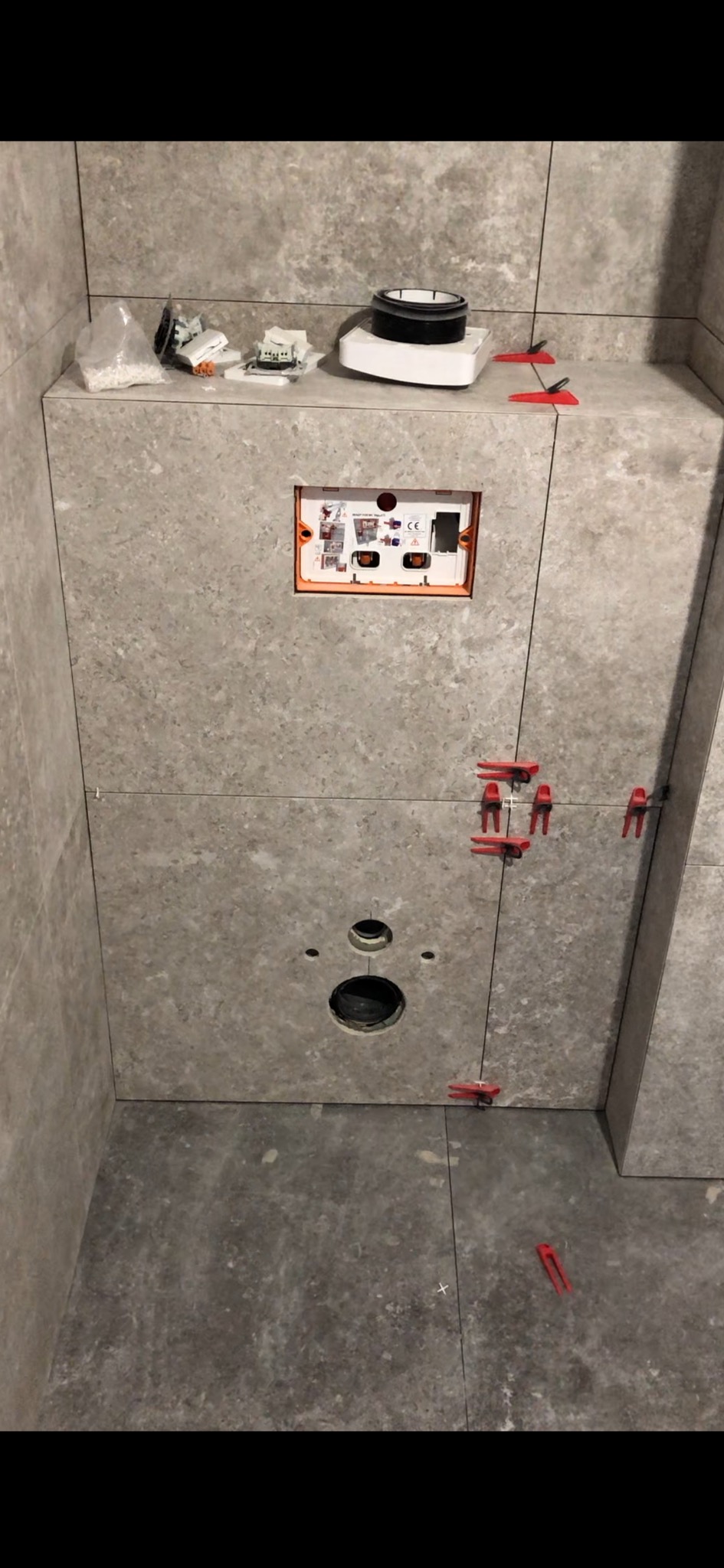 Montaż szarych płytek w łazience, widoczne otwory na instalacje, poziomica laserowa, krzyżyki dystansowe i elementy instalacji podtynkowej.