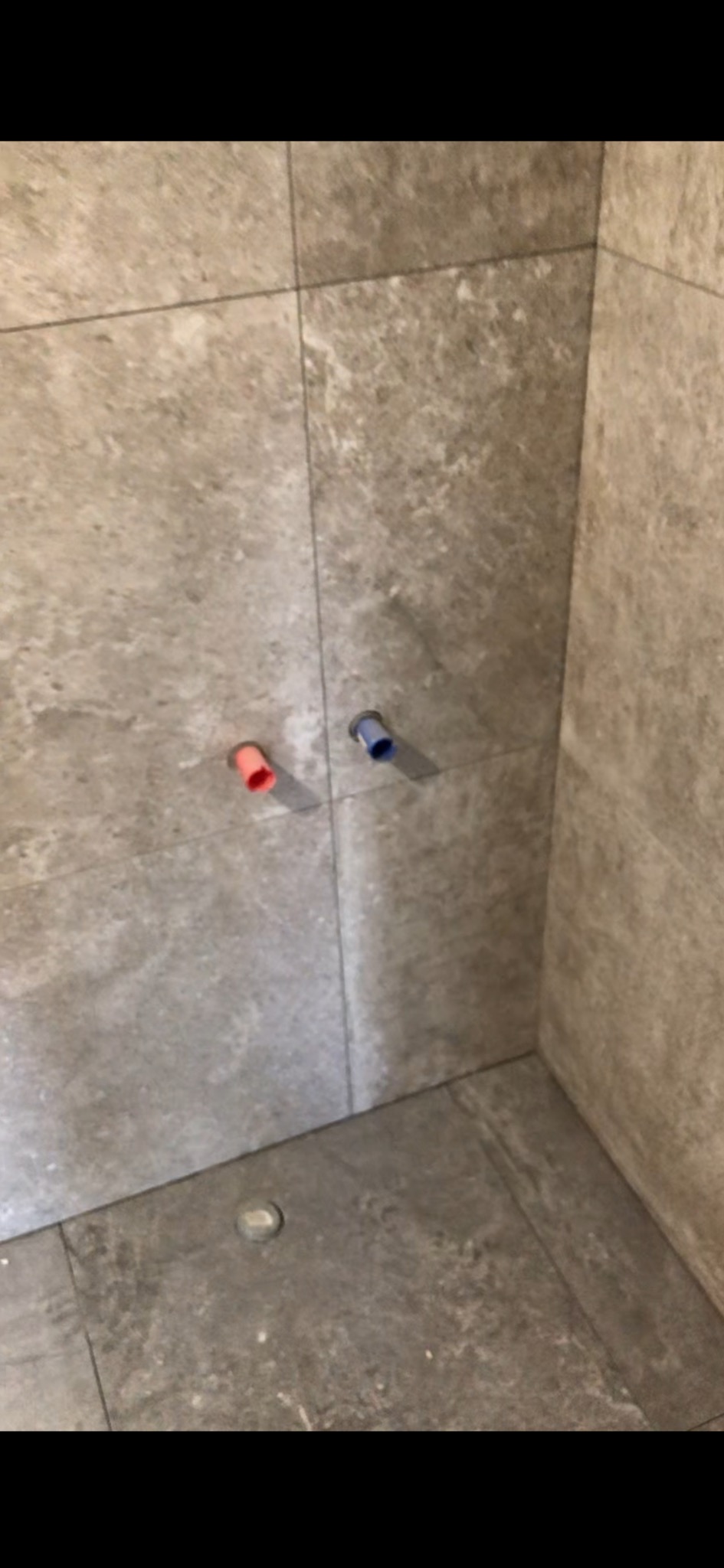 Fragment narożnika łazienki wyłożonego płytkami imitującymi kamień, widoczne wystające rury instalacji wodnej w kolorach czerwonym i niebieskim oraz odpływ podłogowy.