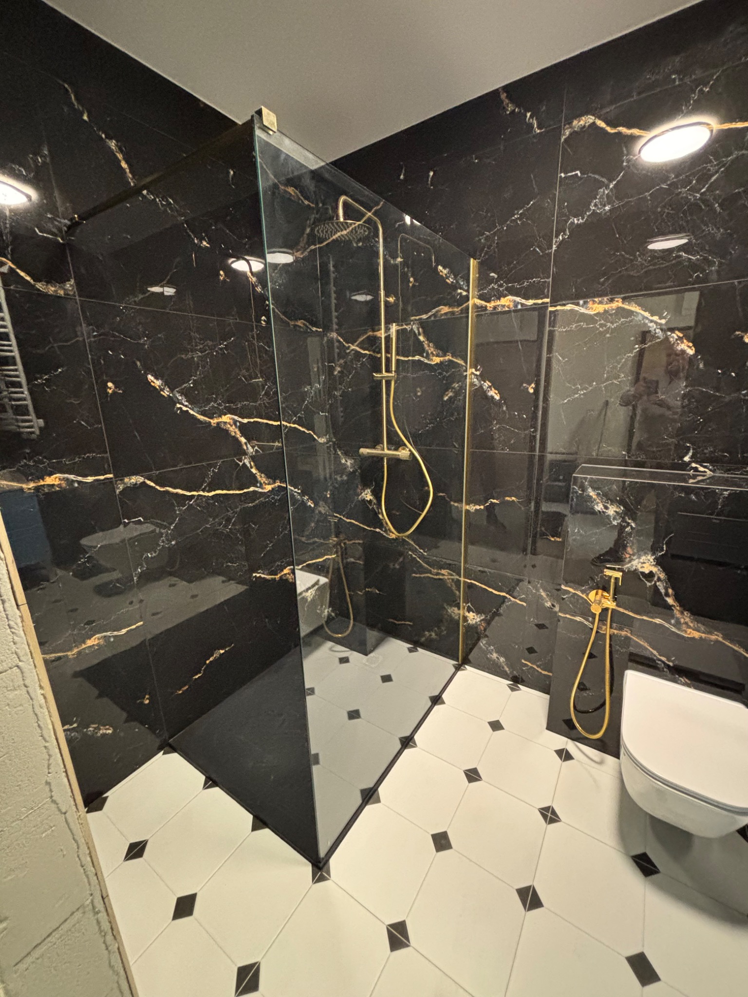 Elegancka łazienka z czarnymi płytkami imitującymi marmur, złotą armaturą i kabiną prysznicową typu walk-in. Biało-czarna podłoga w romby dodaje kontrastu.