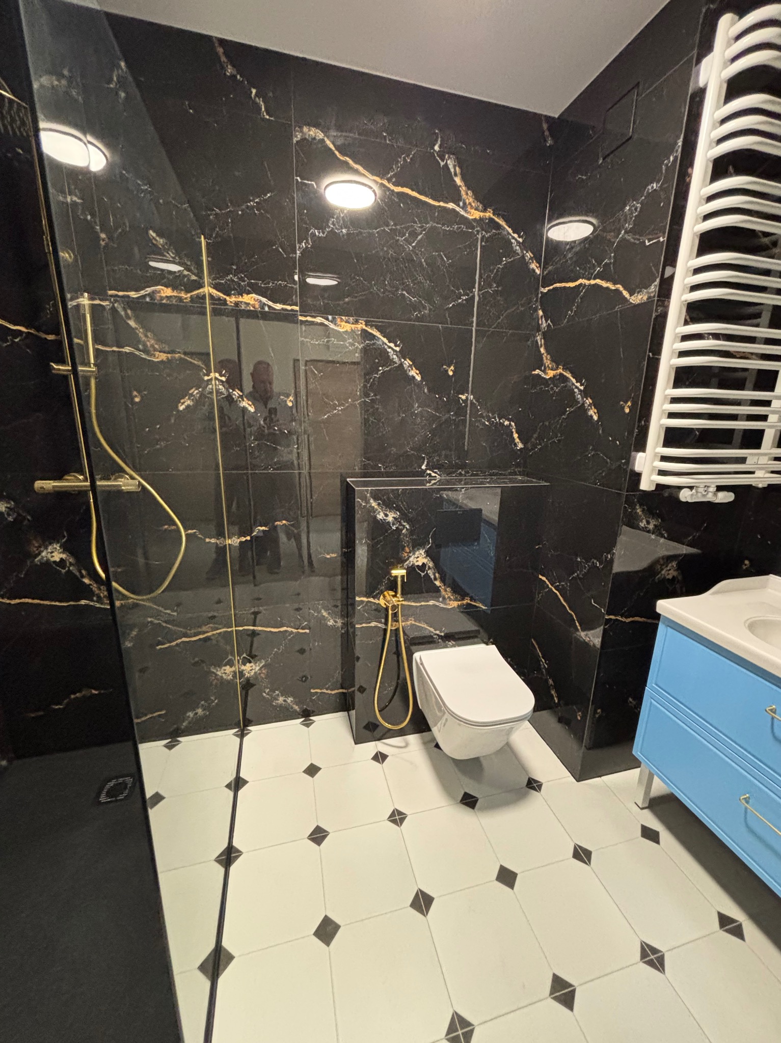 Wykończona łazienka z czarnymi płytkami imitującymi marmur ze złotymi żyłami, białą armaturą i błękitną szafką pod umywalką. Widoczny prysznic i grzejnik.