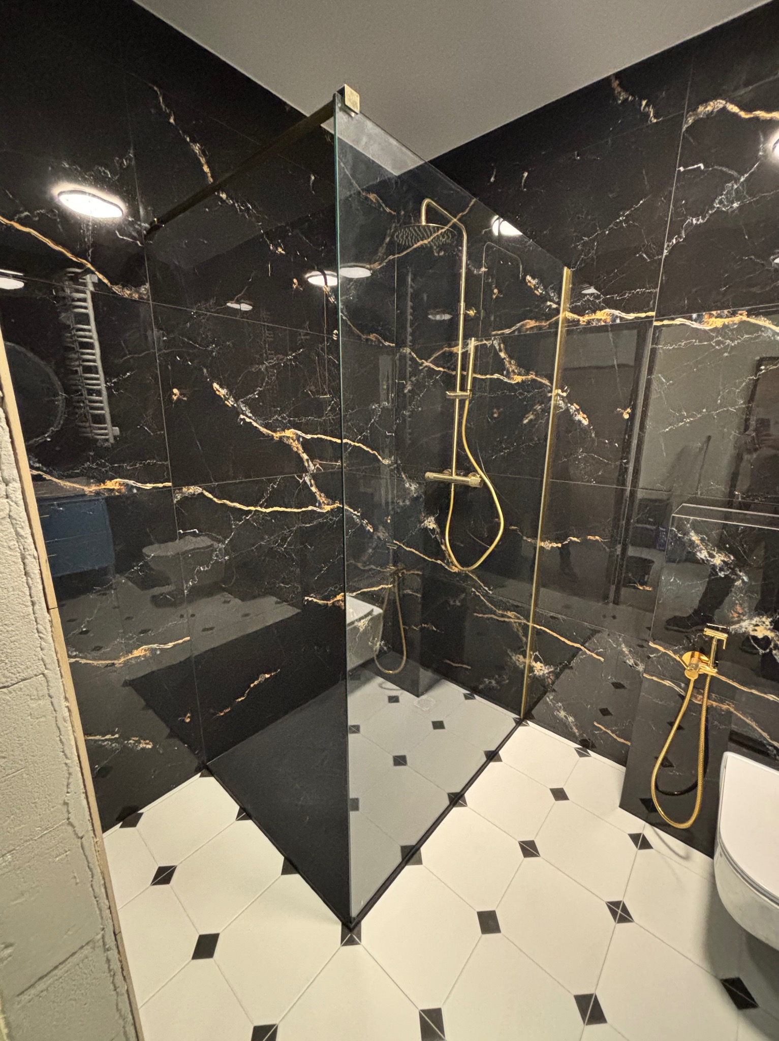 Nowoczesna kabina prysznicowa z czarnymi płytkami imitującymi marmur ze złotymi żyłami i biało-czarną podłogą w romby. Złota armatura.