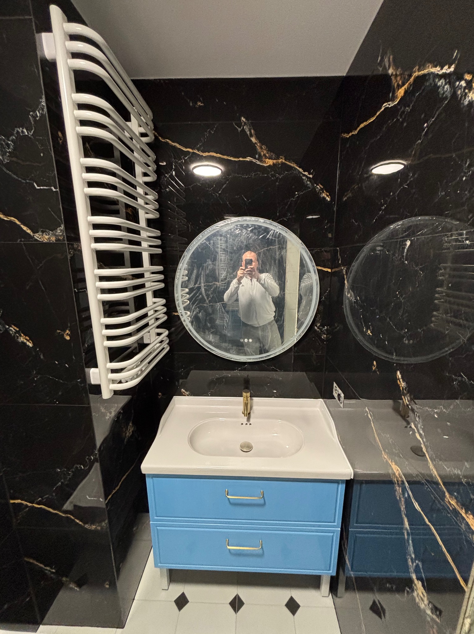 Wykończona łazienka z czarnymi marmurowymi ścianami, błękitną szafką pod umywalką i białym grzejnikiem drabinkowym. W lustrze odbija się osoba robiąca zdjęcie.