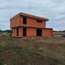 Dwukondygnacyjny dom w trakcie budowy, wzniesiony z czerwonej cegły. Widoczne otwory okienne i drzwiowe, surowy stan. Krajobraz polny w tle, pochmurne niebo.