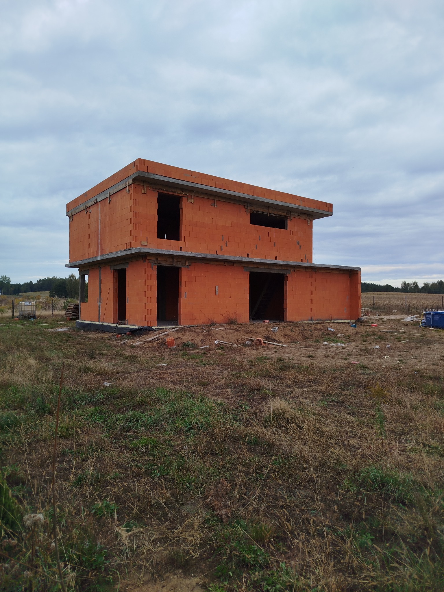 Dwukondygnacyjny dom w trakcie budowy, wzniesiony z czerwonej cegły. Widoczne otwory okienne i drzwiowe, surowy stan. Krajobraz polny w tle, pochmurne niebo.