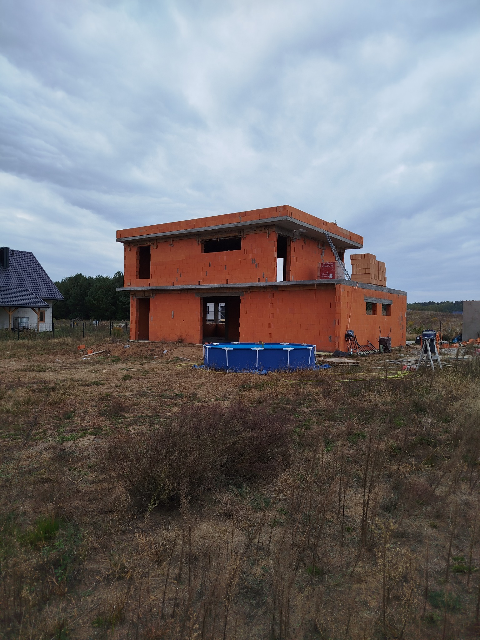 Wznoszony, dwukondygnacyjny dom z czerwonej cegły na działce z widocznym basenem. Częściowo zrealizowana konstrukcja, drabina oparta o ścianę, pochmurne niebo.