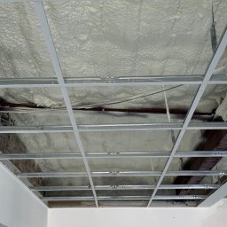 BRC GROUP SPÓŁKA Z OGRANICZONĄ ODPOWIEDZIALNOŚCIĄ - Poddasze ocieplone białą pianą poliuretanową, widoczne metalowe profile konstrukcyjne sufitu podwieszanego i fragment ściany z cegły