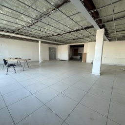 BRC GROUP SPÓŁKA Z OGRANICZONĄ ODPOWIEDZIALNOŚCIĄ - Wnętrze dużego pomieszczenia z widoczną konstrukcją metalową pod sufitem wypełnioną jasną pianą izolacyjną, białe ściany z widocznymi śladami po pracach remontowych, biała podłoga z płytek, stolik...