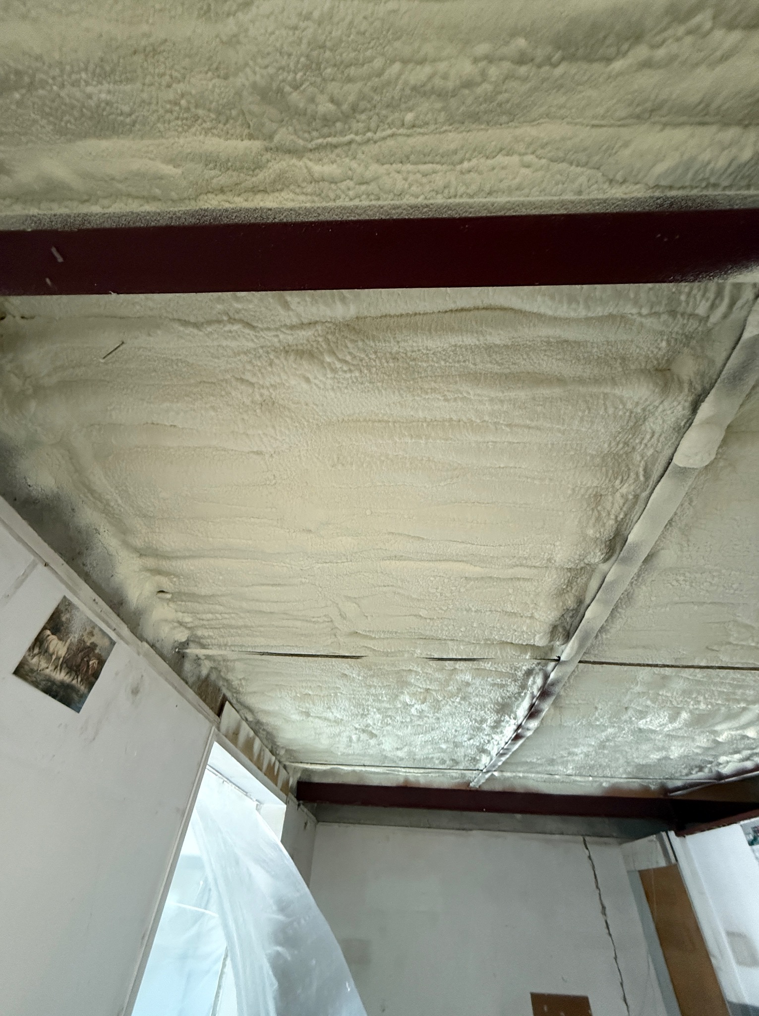 Poddasze z widoczną strukturą natryśniętej, jasnej pianki izolacyjnej, przylegającej do metalowych belek konstrukcyjnych; po lewej stronie ściana z wiszącym obrazkiem.