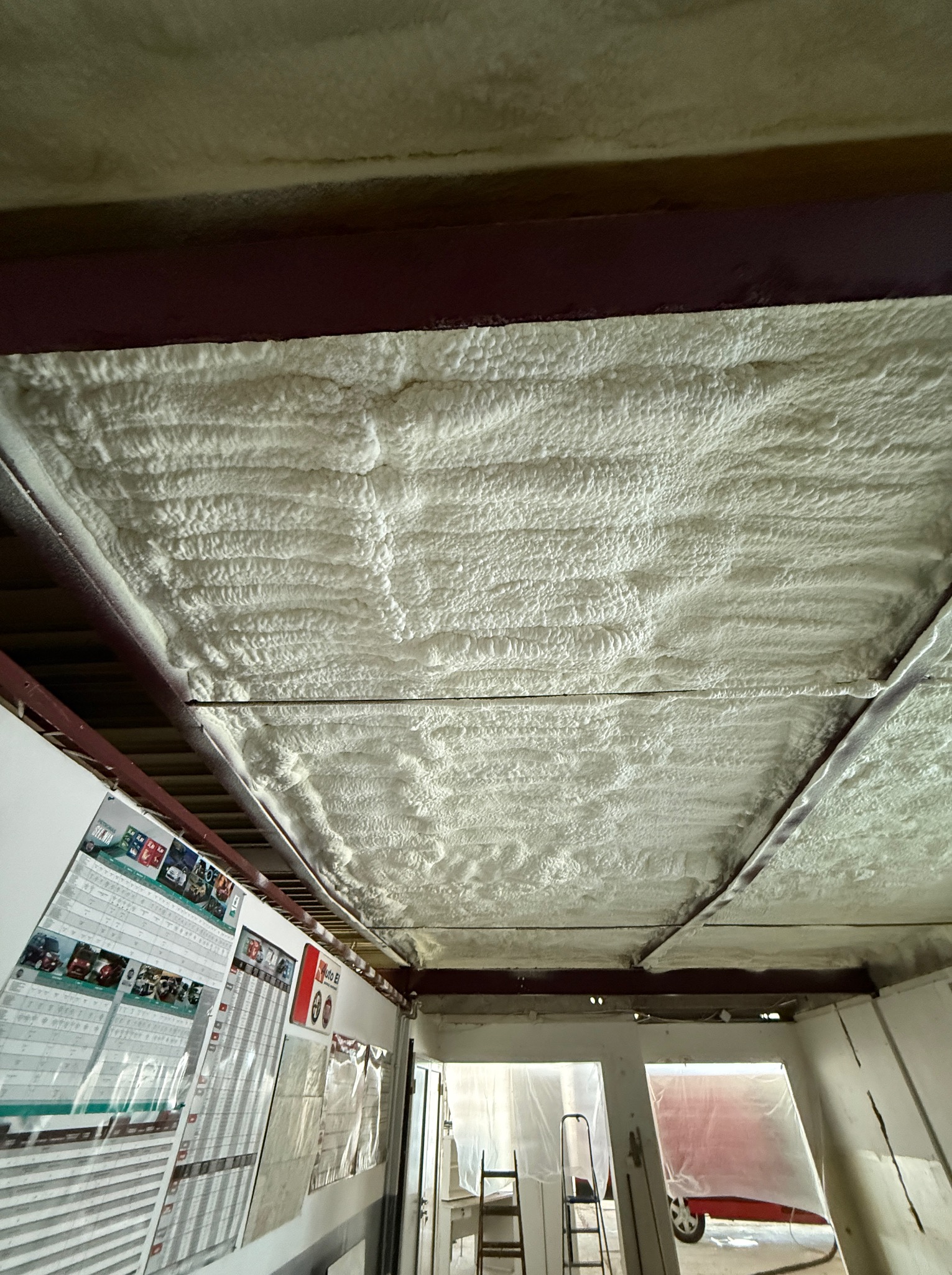 Poddasze zabezpieczone grubą warstwą izolacji natryskowej z pianki, widoczne stalowe belki konstrukcyjne, w tle plakaty i pomieszczenie w trakcie remontu.