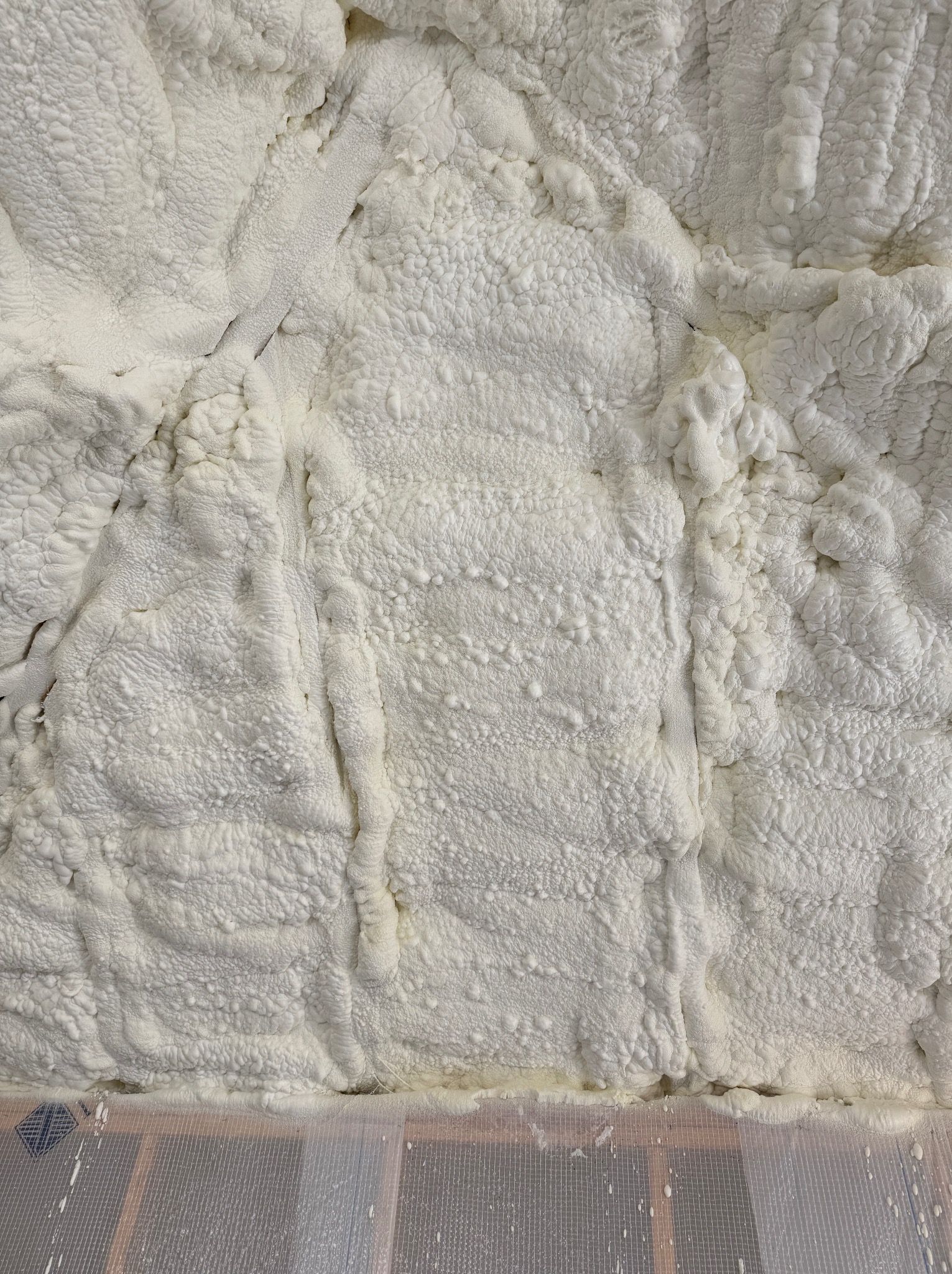 Świeżo natryśnięta, jasna piana izolacyjna wypełnia przestrzeń między elementami konstrukcji drewnianej, tworząc nierówną, porowatą powierzchnię o gęstej strukturze.