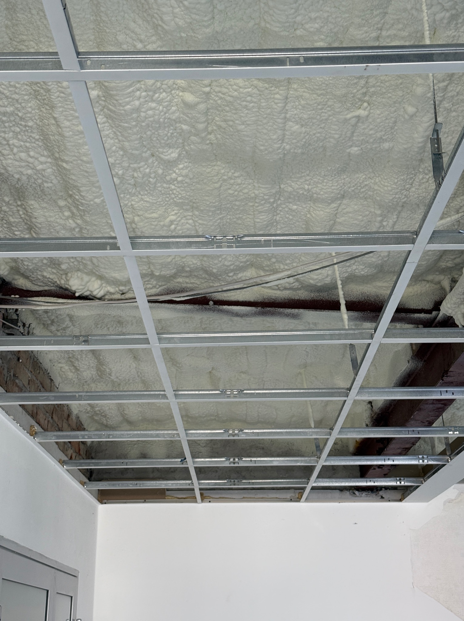 Poddasze ocieplone białą pianą poliuretanową, widoczne metalowe profile konstrukcyjne sufitu podwieszanego i fragment ściany z cegły