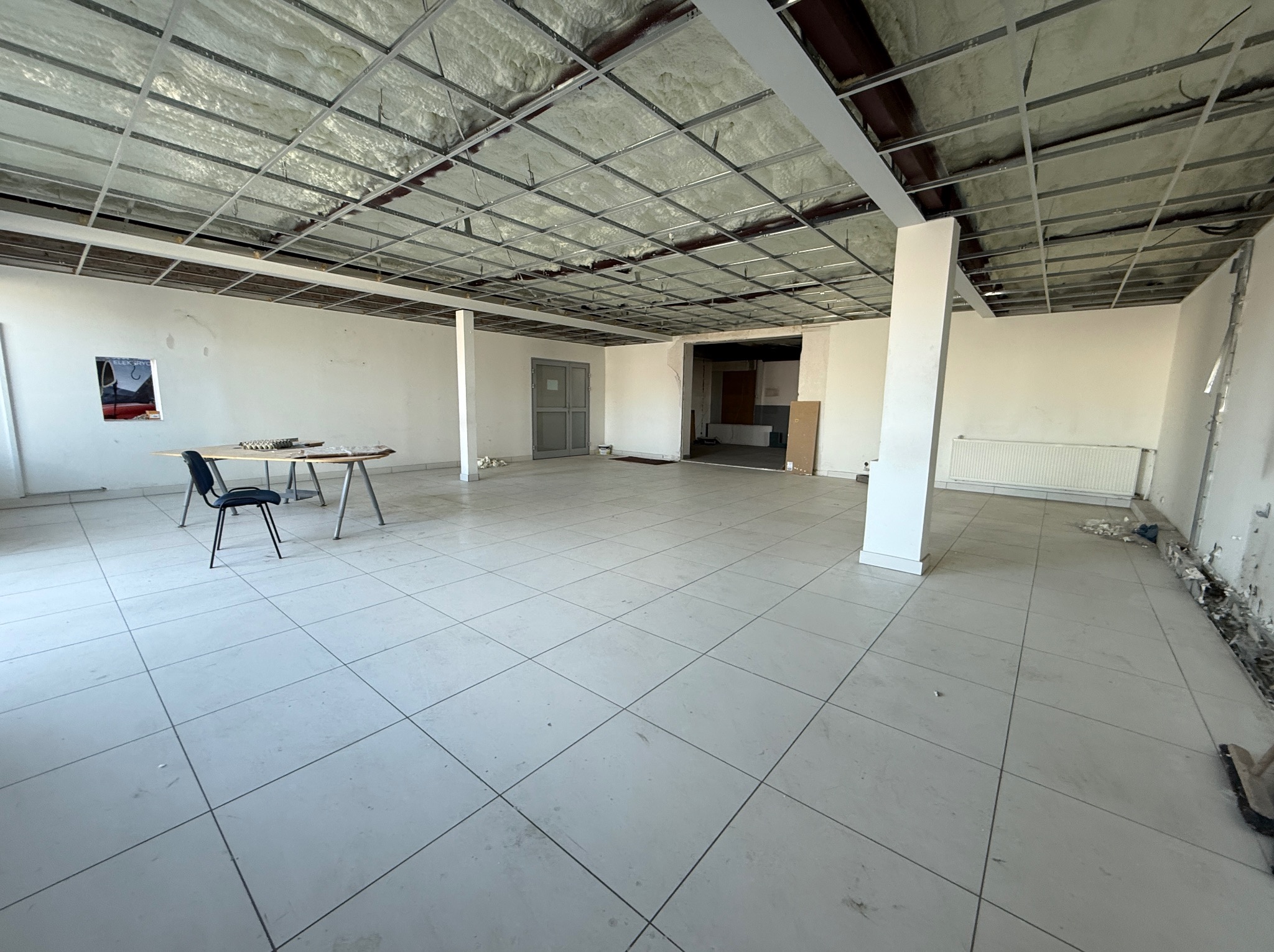 Wnętrze dużego pomieszczenia z widoczną konstrukcją metalową pod sufitem wypełnioną jasną pianą izolacyjną, białe ściany z widocznymi śladami po pracach remontowych, biała podłoga z płytek, stolik...