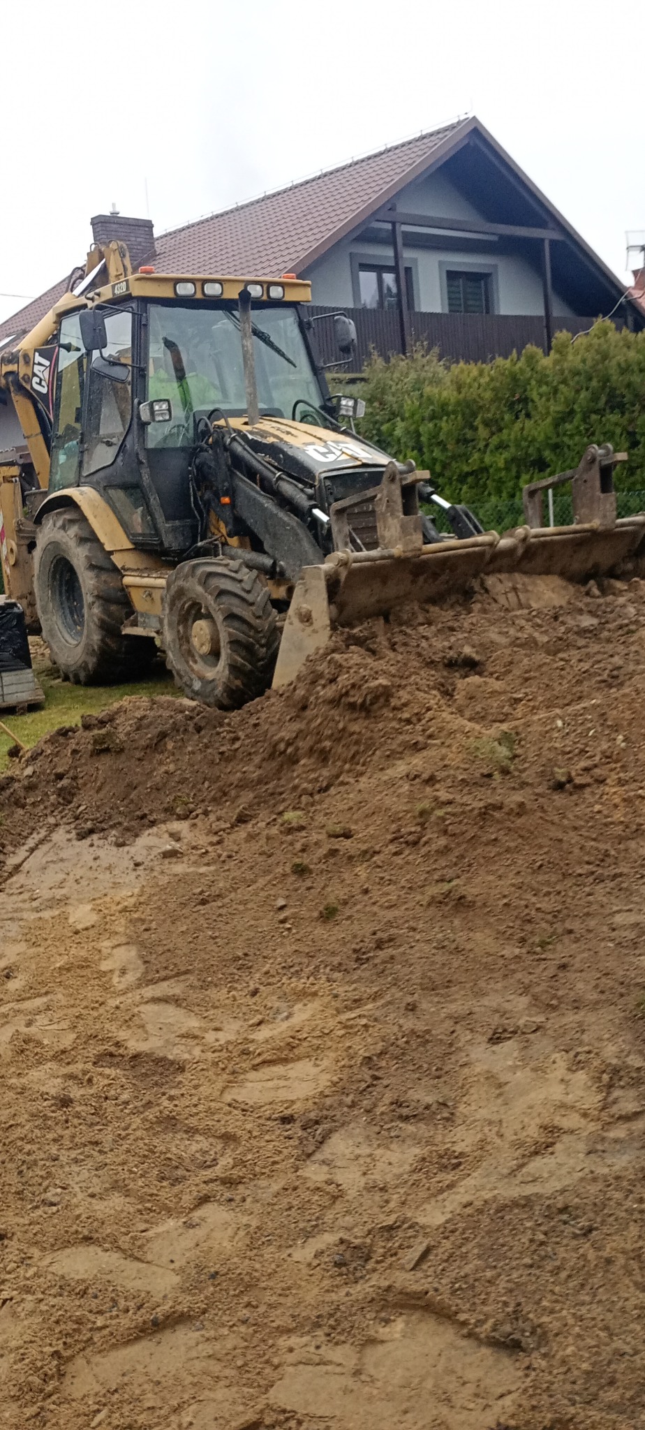 Żółto-czarna koparko-ładowarka CAT nabiera brudną ziemię na tle domu z brązowym dachem.