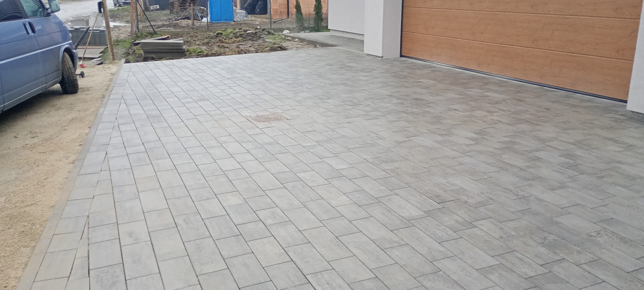 Nowo ułożona, szara kostka brukowa prowadząca do garażu, z widoczną studzienką kanalizacyjną i samochodem dostawczym w tle.