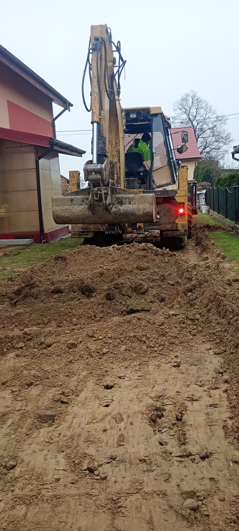 Żółta koparko-ładowarka w trakcie pracy na działce, widoczne świeżo naruszone podłoże i ślady opon, operator w kabinie, budynek w tle.