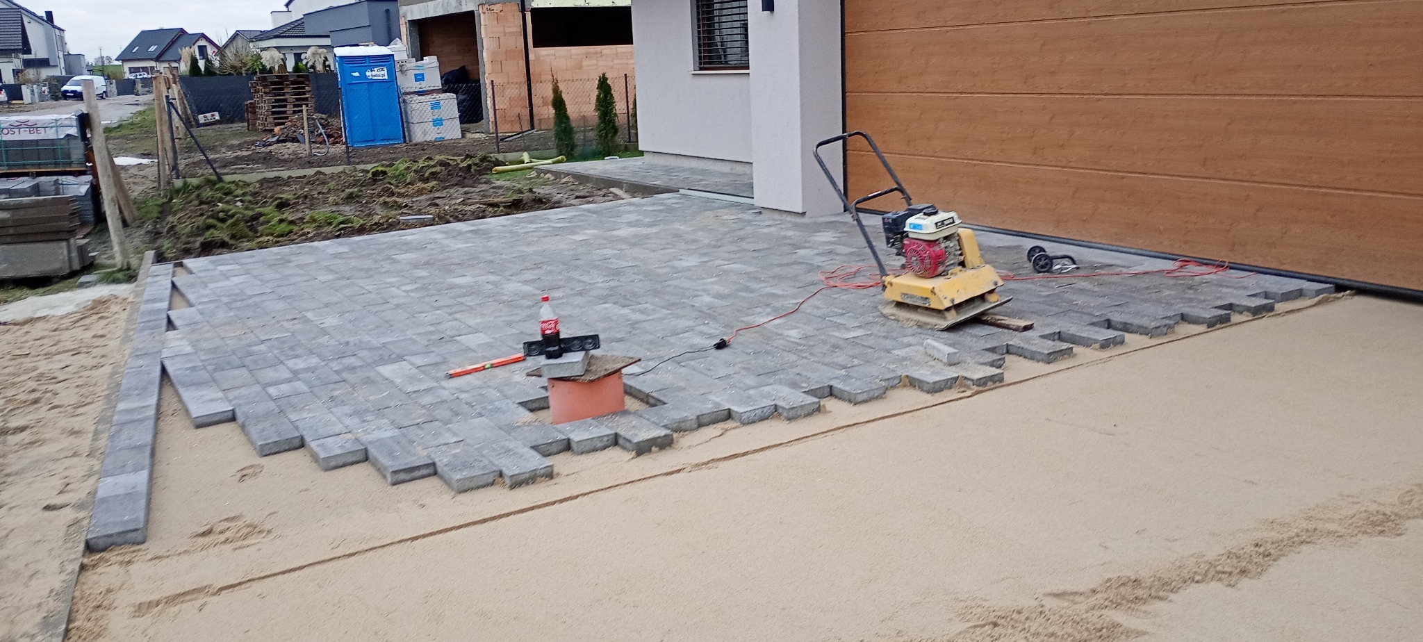 Fragment układanej szarej kostki brukowej na podsypce piaskowej, widoczna zagęszczarka wibracyjna, poziomica, butelka Coca-Coli na prowizorycznym podeście z rury PCV i głośnika, w tle budynek...