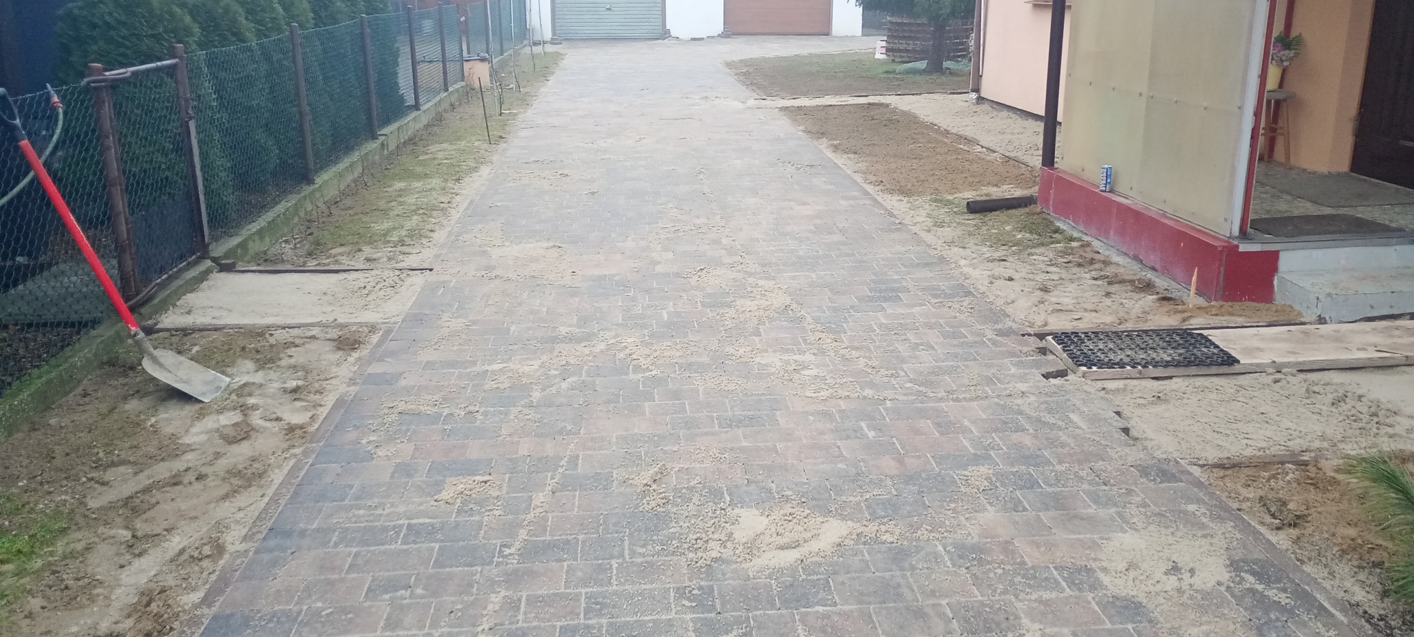 Nowa, częściowo wyłożona kostka brukowa na podjeździe, z widoczną łopatą opartą o ogrodzenie i piaskiem rozsypanym na powierzchni.
