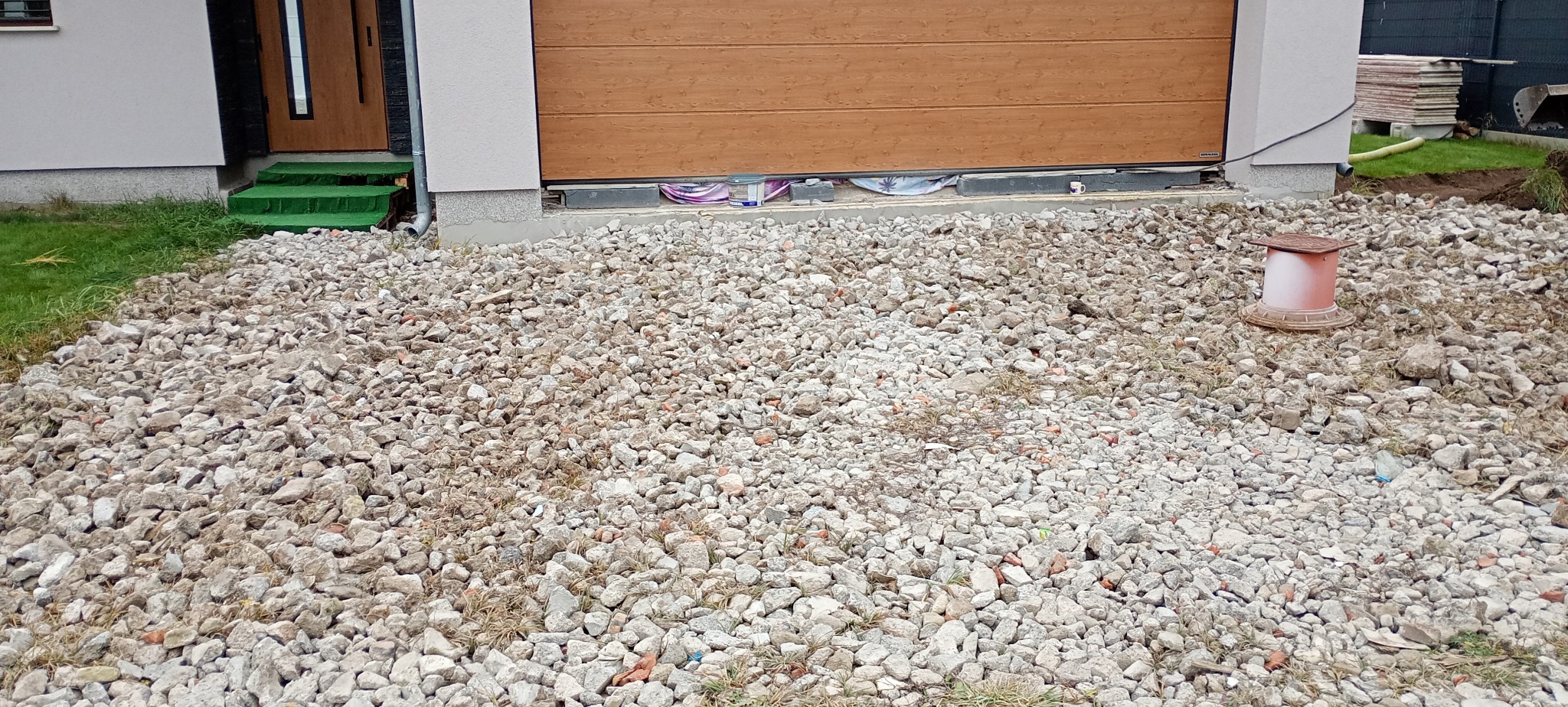 Teren przed budynkiem z wysypanym gruzem i kamieniami, przygotowywany pod utwardzenie drogi, widoczna brama garażowa i wejście do domu z zielonymi schodami.