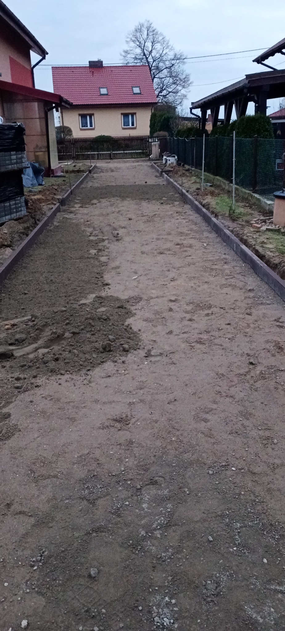 Przygotowany teren pod ścieżkę z obrzeżami, częściowo zasypany piaskiem, widoczne budynki w tle.