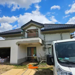 Dom w trakcie ocieplania: elewacja częściowo otynkowana, rusztowania, zakryte okna, wiaderka z farbami w garażu, samochód dostawczy z przodu.