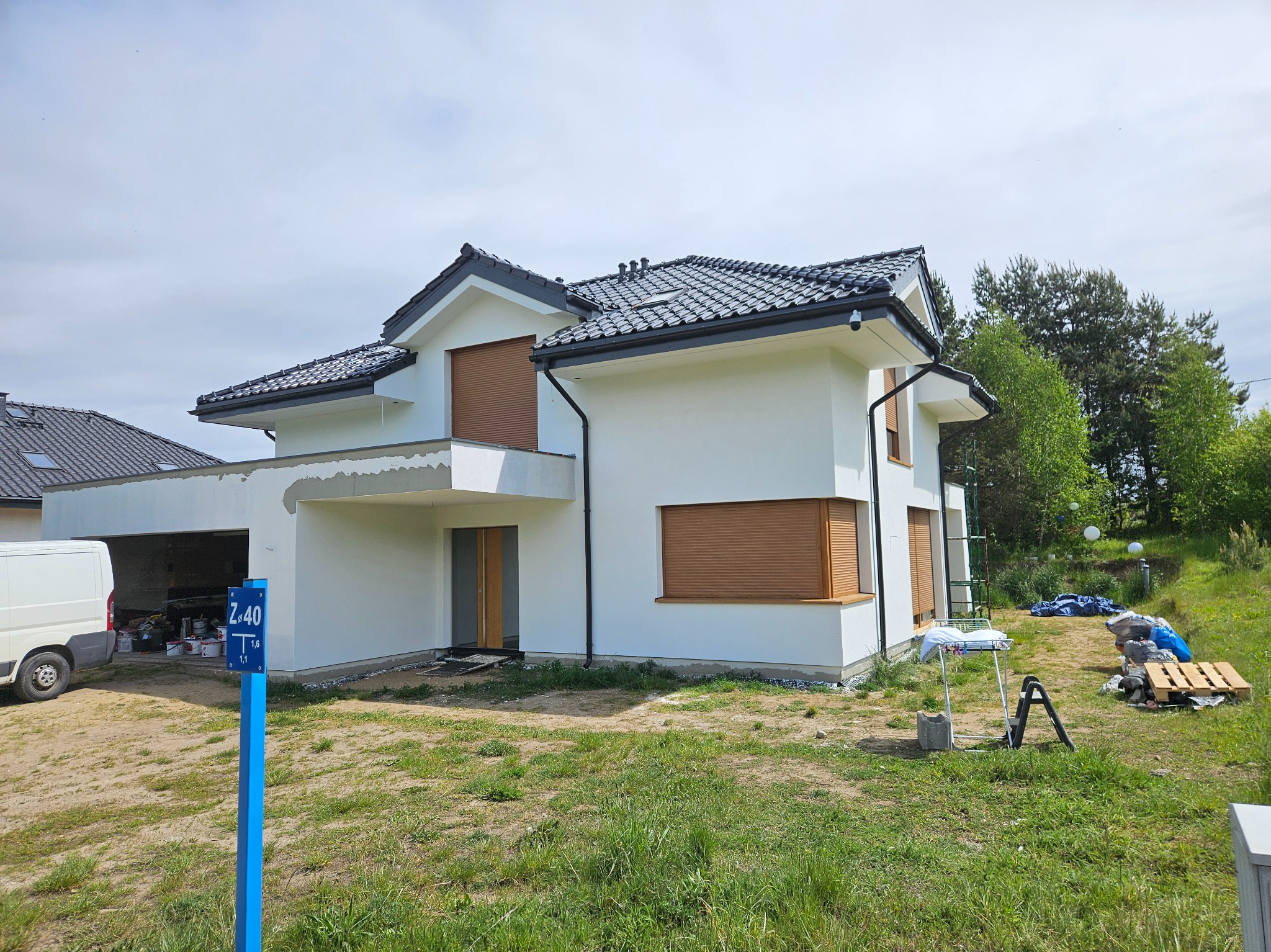 Biały, dwukondygnacyjny dom z brązowymi roletami i czarnym dachem, widoczny z zewnątrz, z elementami wykończeniowymi. Na trawniku narzędzia i materiały budowlane.