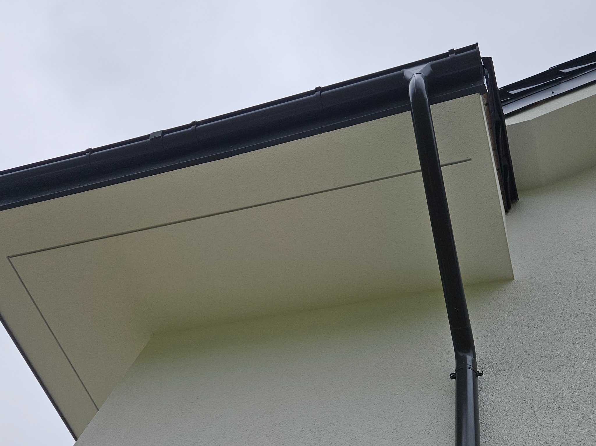 Fragment elewacji budynku z zamontowaną, ciemną rynną spustową na tle jasnej ściany z fakturą i pochmurnego nieba. Widok z dołu.